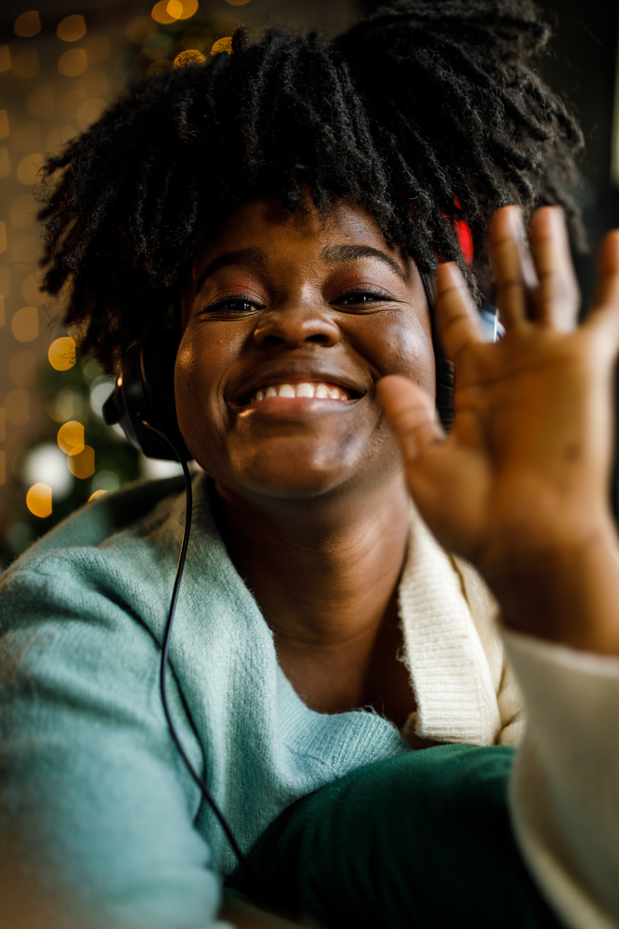 Joyful young Black woman wearing headphones, waving and smiling at camera as if on a video call